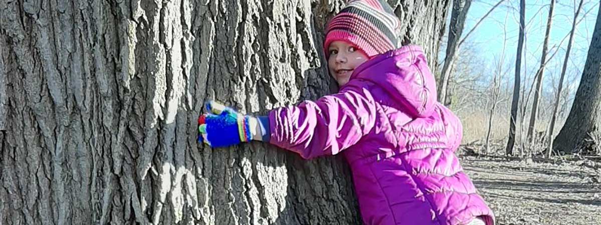 child in winter jacket, hat and gloves hugging a very large tree