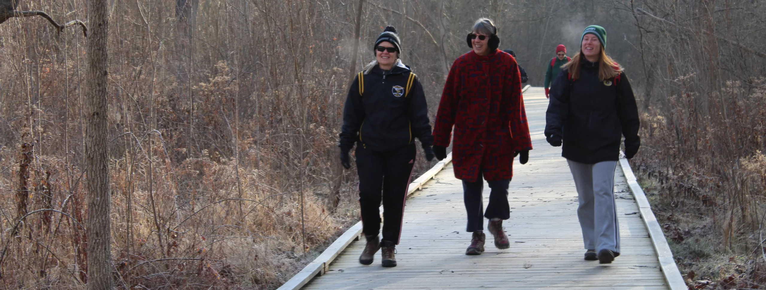 Three people smile while walking down boardwalk trail surrounded by woods during winter. They are bundled in jackets and hats. Two more people walk a distance behind them.