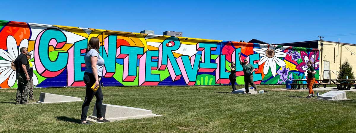 A group of people playing cornhole in front of a colorful mural that says Centerville