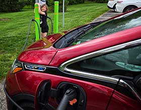 A red electric vehicle hooked up to an EV chargins station in a public park. A young girl watches in the background.