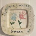 white plate painted with children's handprints to look like flowers with the words papa's and ganma's garden written in black around the edge