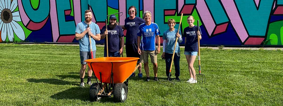 A group of people standing outside with rakes. An orange wheelbarrow is in front of them.