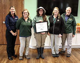 Five people standing in a room. The person in the middle holds a framed document.