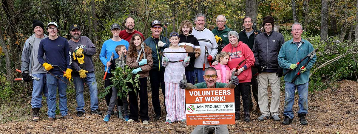 A group of people standing outside. One man is giving two thumbs up while kneeling behind a sign that says "Volunteers at Work!"