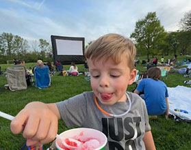 Young boy with tongue sticking out scoops frozen ice dessert with a spoon. There is a inflatable movie screen in the background.