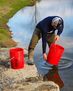A man with two red buckets with one foot in a pond and the other foot on a rock at the pond's edge.