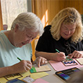 Image of two adults completing a craft while sitting next to each other. Both are using markers to decorate their craft.