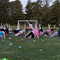 Image of multiple individuals participating in yoga in the evening. They are in a side plank with an arm stretched into the air. The instructor stands in the center of the image. Colorful round orb lights are in the grass.