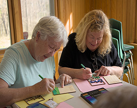 Image of two adults completing a craft while sitting next to each other. Both are using markers to decorate their craft.