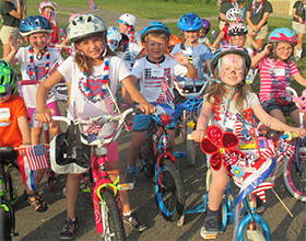 A large group of children on bicycles and wearing helmets. Many have decorated their bikes with patriotic or red, white and blue decorations.