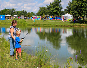 An adult and a child stand on the edge of a pond. The child holds a fishing pole. There are colorful inflatables and tents at the event on the other side of the pond.