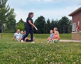 A person is standing in the grass. Several people are seated around her.