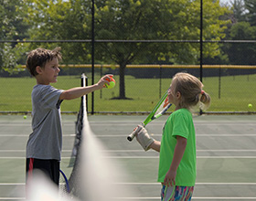 Two young children stand on either side of a tennis net. One holds a racquet and the other hands them a red and green tennis ball over the net.