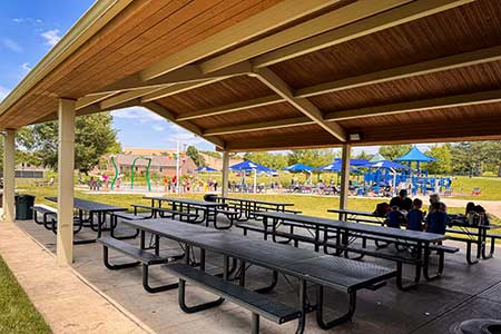 The inside of a picnic shelter. One table is in use by a family. Splash pad and playground can be seen in the background.