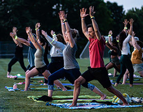 Image of multiple people participating in an evening outdoor yoga class in a park grassy field. They stand on towels and yoga mats. Everyone is standing in a warrior pose, lunging with one knee and arms stretched up above their heads. Everyone wears glow bracelets.