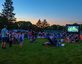 A group of people sit on blankets or in lawn chairs in a field watching a movie on an inflatable screen at sunset.