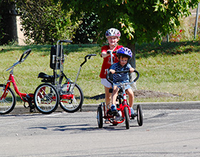 Image of two children in helmets and adapted bikes. A younger child in blue pedals the bike and an older child pushes the bike from behind.