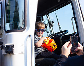 Image of a young child sitting in a large truck while holding an orange Halloween candy bucket and wearing sunglasses. An adult uses a cellphone to take a picture of the child.
