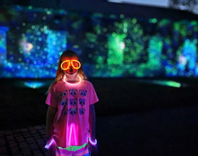 Image of a child in a bright pink shirt where glow in the dark glasses and necklace. The background is mostly dark with some blue and green light illuminating a brick wall.