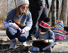 An adult wearing a beanie hat, sunglasses, and a shirt that reads "The Great Turkey Trek" squats next to a child wearing a matching shirt and a red and back beaning who is coloring on a piece of paper on a clipboard while sitting crisscrossed. Another adult and child are in the background.
