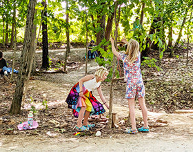 Image of two children building a fairy house in the woods. One wears a colorful, butterfly-inspired skirt. The other wears a floral print outfit. One child squats to add to the fairy house, the other is tying string to a branch of a tree.